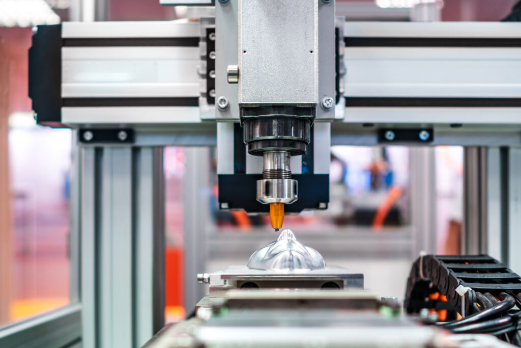 close-up view of a modern industrial 3D printer or CNC machine, actively working on a metallic object. The machine's head is precise, with a nozzle or cutting tool, and the object being crafted appears to have a smooth, reflective surface. The background is slightly blurred, showcasing an industrial environment.