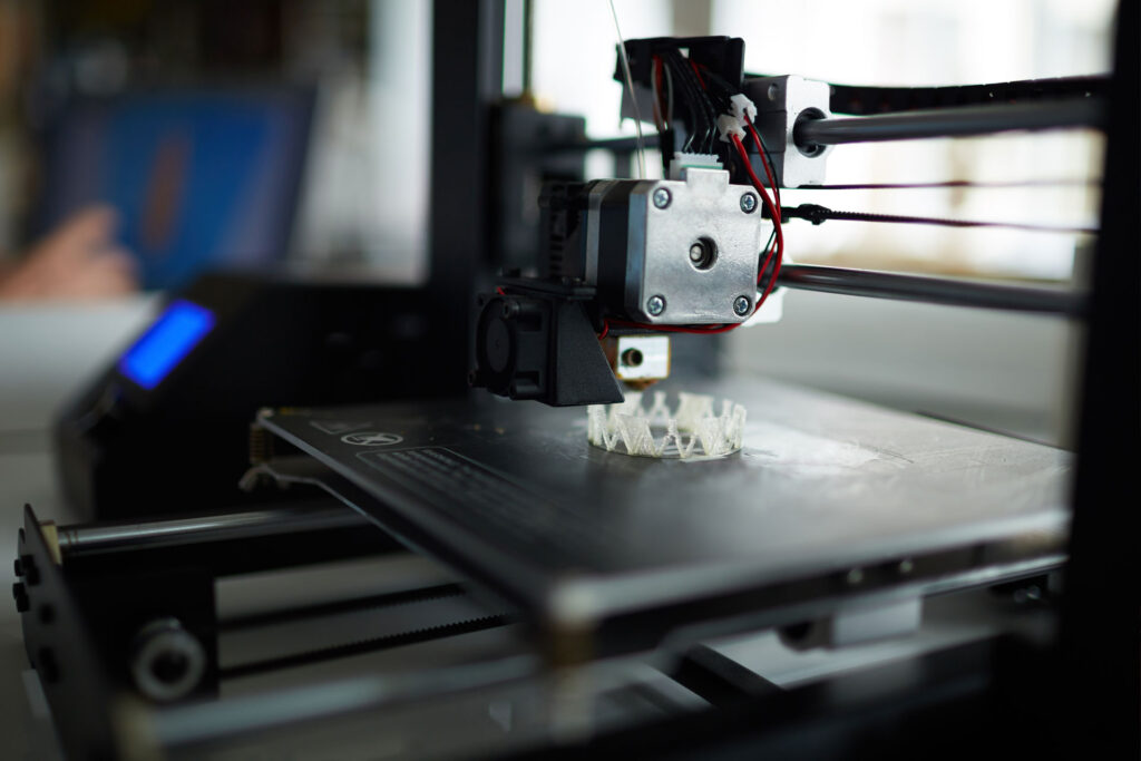 Close-up view of a 3D printing machine in operation, creating a small intricate object layer by layer. The print head, wiring, and partially completed design emphasize precision and modern manufacturing technology.