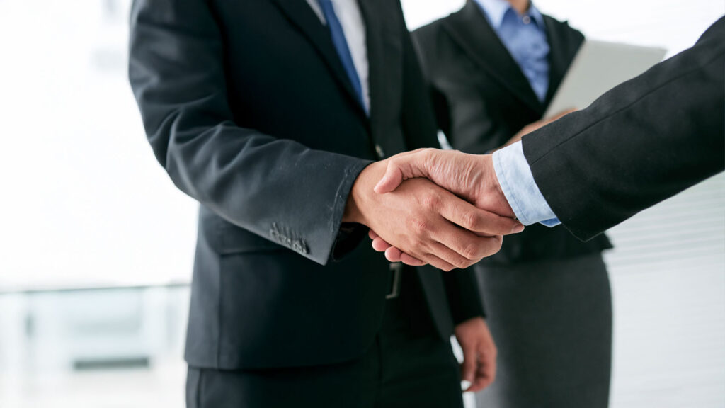 Close-up of a handshake between two individuals in formal business attire, symbolizing a professional agreement or partnership, with a third person holding documents in the background.